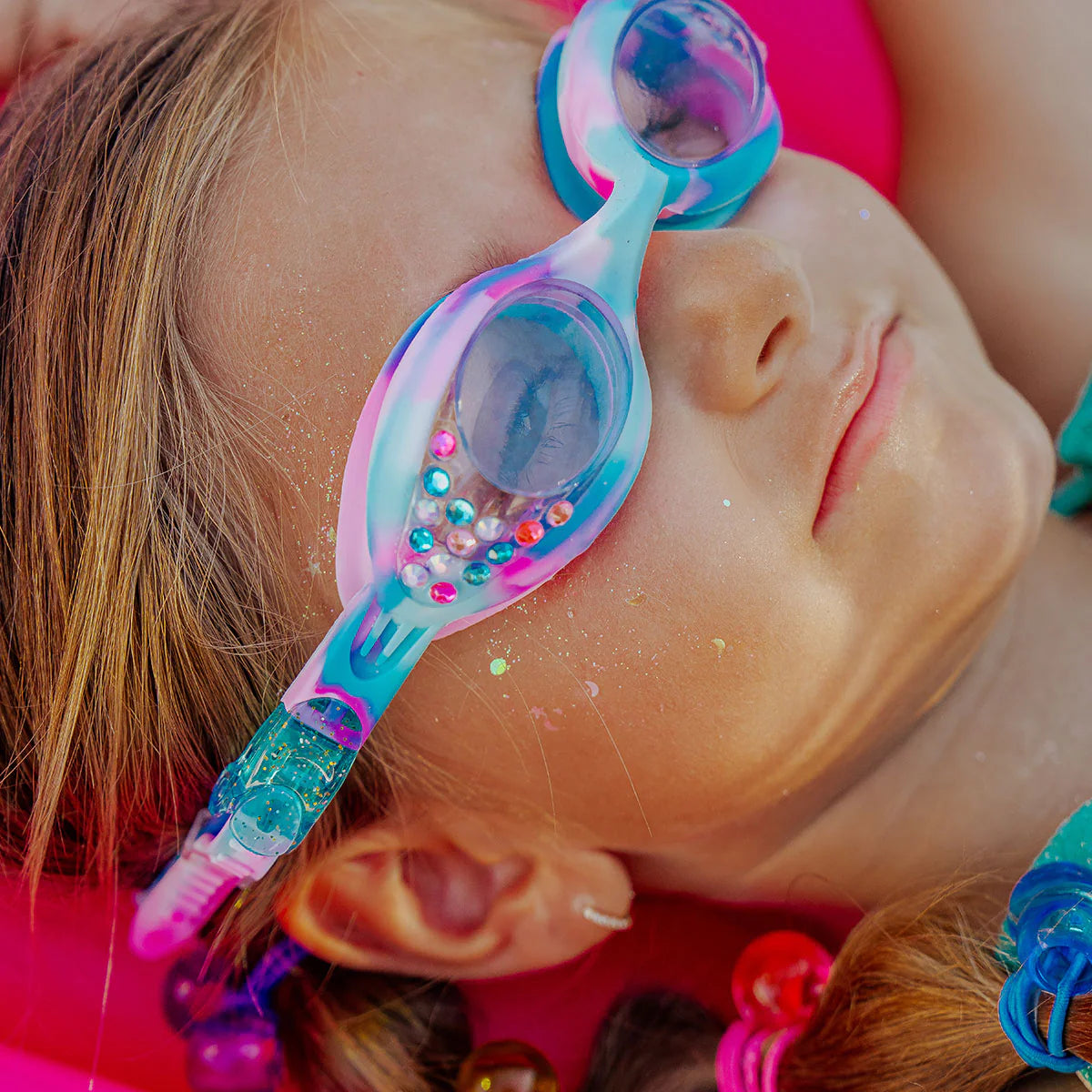 A child with glitter on their face wears Bling2O Tiny Taffy Swim Goggles, decorated with beads and rhinestones, while lying down and facing upward.