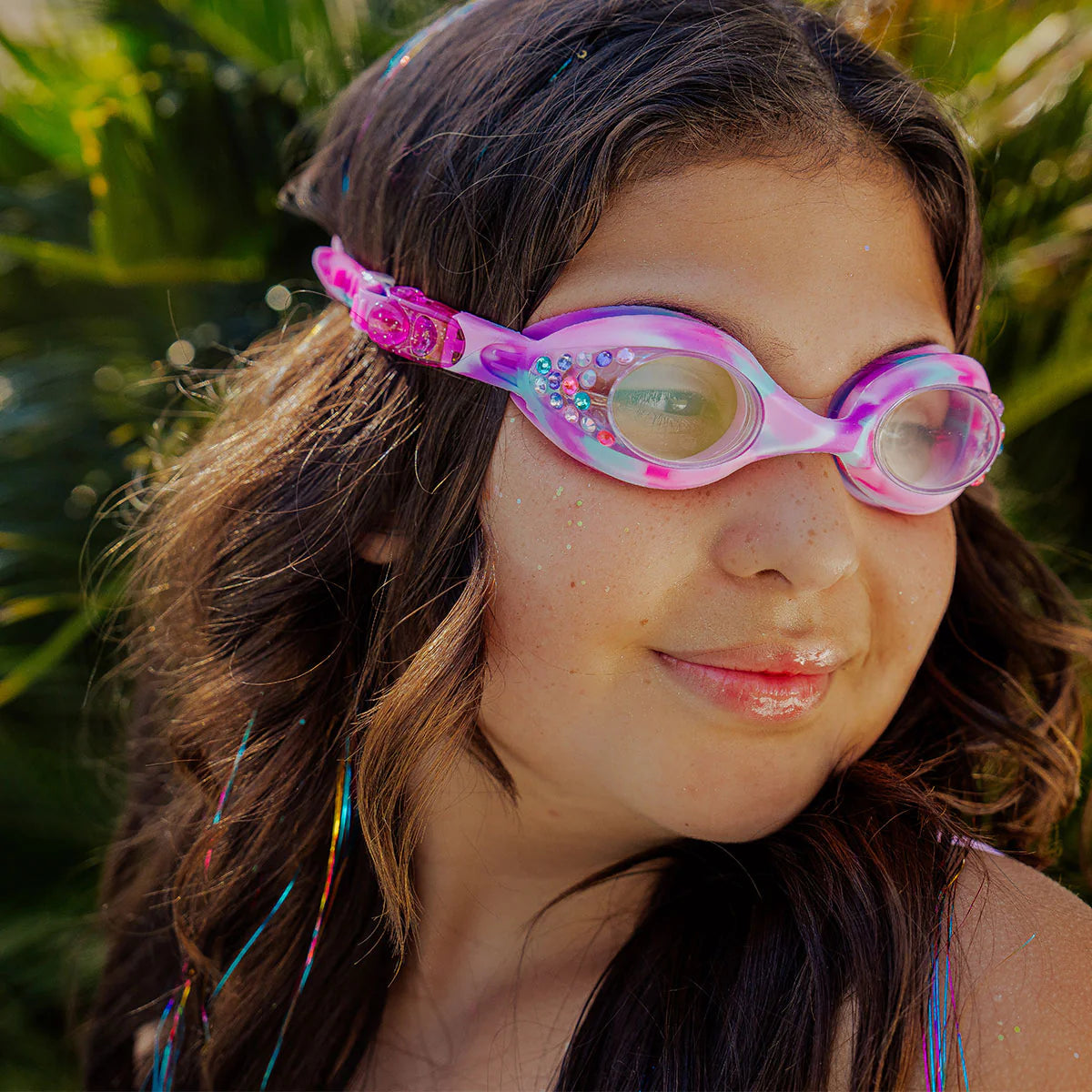 A girl with long brown hair stands outdoors in front of green foliage, looking to the side while wearing pink Tiny Taffy Swim Goggles from Bling2O.