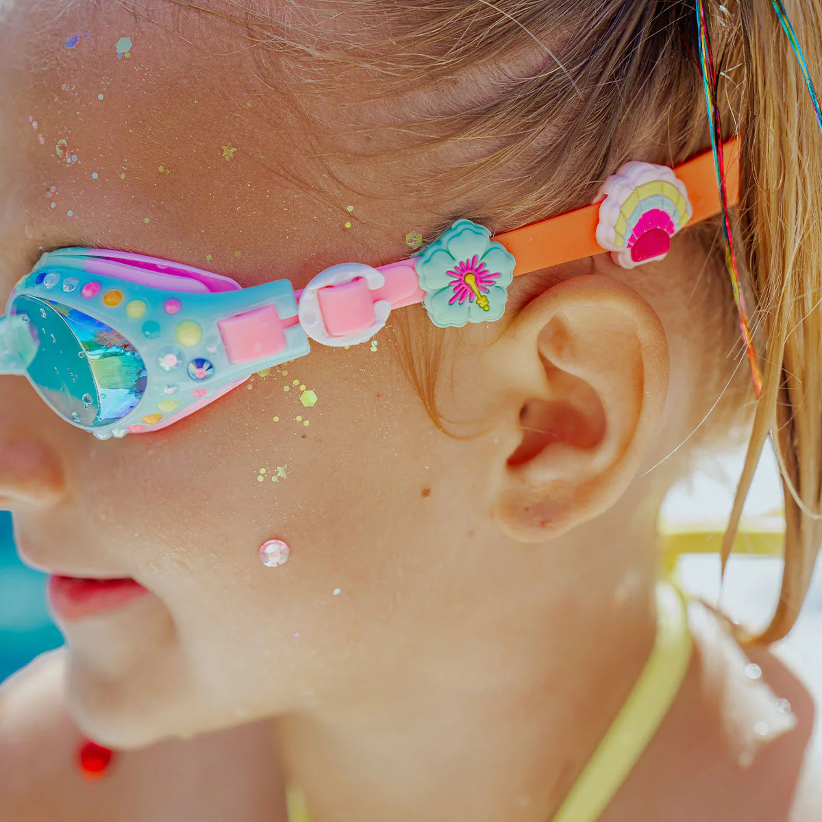 Close-up of a child wearing Bling2O Tropical Rhinestone Swim Goggles, featuring beads, a flower, rainbow charm, and sparkling rhinestones, with glitter decorating their face and hair.