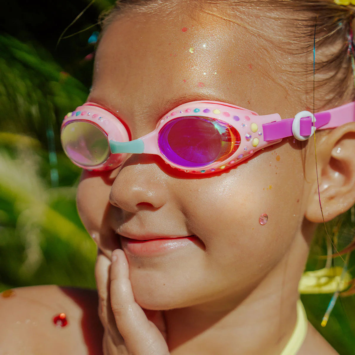 A young child wearing Bling2O's Tropical Rhinestone Swim Goggles smiles and touches their chin, with sunshine and greenery in the background.