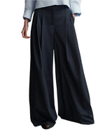 A person is casually posing with hands in pockets, wearing TWP New Didi Wool Pants, elegant pleated wide-leg black trousers made from Italian Wool Twill, paired with a light blue shirt.