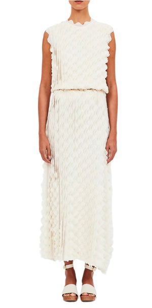 The individual is dressed in a sleek Ulla Johnson Kaelyn Dress, adorned with scalloped edges and a sleeveless textured design in pristine white, paired perfectly with metallic sandals.