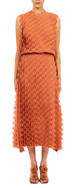 A person wears the Ulla Johnson Kaelyn Dress, an orange scallop ruffle knit dress with a wavy pattern. The sleeveless nylon top transitions into a mid-length skirt. Matching orange shoes complete the outfit, as they stand with arms by their sides.