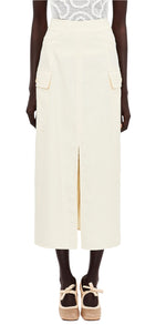 An individual is dressed in a white patterned top and an Ulla Johnson Rhodes Skirt, a cream-colored cotton tencel piece with pockets and front slit. They complement it with beige platform shoes featuring ankle straps.