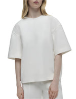 A person is wearing the Simkhai Aude Top, a loose-fitting white short sleeve top paired with white pants crafted from heavy cotton. Against a plain background, this minimalist ensemble by Simkhai highlights elegance and simplicity with its refined silhouette.