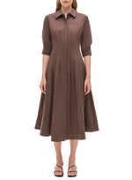 A woman wears the Simkhai Signature Jazz Dress—a brown cotton poplin midi with an A-line silhouette, collared neckline, elbow sleeves, and pleated skirt—paired with black open-toe sandals.