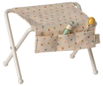 The Maileg Nursery Table, Baby Mouse - Off White, features a beige polka-dot seat with pockets to hold accessories like a baby bottle, yellow pacifier, and tiny rattle—ideal for a cozy baby mice nursery setup.