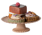 The Maileg Cakes and Cakestand, Mini features an elegant assortment of mini cakes, including a square chocolate cake and round tarts, beautifully displayed on a fluted beige stand with berry and floral garnishes.
