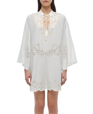 A woman wears the Simkhai Sarina Mini Dress—a white, long-sleeve cover-up with scalloped edges, broderie anglaise, eyelet cutouts, and a tie neckline—perfectly lightweight for warm days.