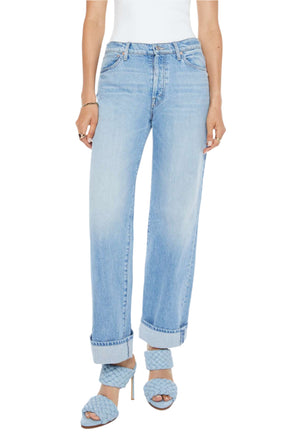 A person stands against a plain background wearing Mother’s Dodger Button Hover Cuff mid-blue denim loose straight leg jeans, a white sleeveless top, and blue braided heeled sandals.
