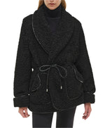 A person wearing the Simkhai Kimia Tie Waist Jacket—a black textured coat with a foldover collar, large lapels, front pockets, cuffed sleeves—paired with a black turtleneck and pants.