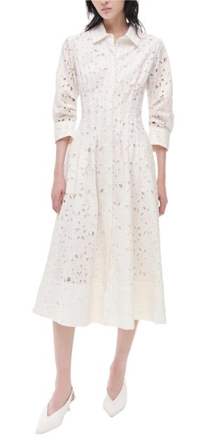 A woman exudes classic femininity in the Simkhai Jazz Floral Broderie Anglaise Dress—a white, long-sleeved collared midi with floral lace details and a fitted waist—paired with chic white pointed-toe heels.
