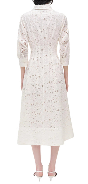 A person shown from the back wears Simkhai’s Jazz Floral Broderie Anglaise Dress—a white, long-sleeved, calf-length lace dress with a fitted waist—paired with white heeled sandals, exuding classic femininity through delicate floral details.
