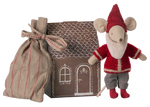 The Maileg Santa Mouse wears a red outfit and hat, standing beside a striped cloth bag and a cardboard Mouse Gingerbread House featuring white door and window designs.