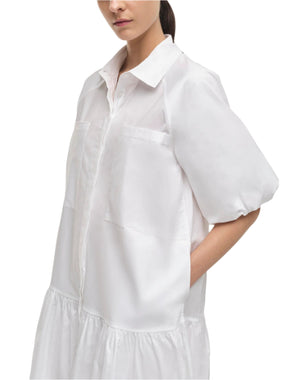 A woman wears the Simkhai Crissy Dress, a loose-fitting white mini dress with a shirting style, puffed sleeves, front and side pockets, and her hands in the side pockets.