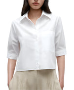 A woman wears the TWP Short Lived Top—a white, boxy fit, short-sleeved cotton poplin button-up shirt with a chest pocket—paired with beige pleated pants, shown from shoulders to hips.