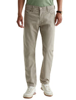 A person is shown from shoulders to feet against a plain background, wearing AG Jeans Tellis Commuter Performance Airluxe Modern Slim pants in light beige, paired with a green shirt, white sneakers, and a wristwatch.