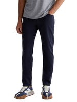 A person stands wearing a gray t-shirt, AG Jeans' Tellis Modern Slim Pant in Italian sateen dark blue, and navy and white sneakers with brown soles.