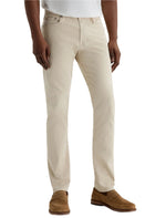 A person wearing AG Jeans Tellis Modern Slim Sueded Stretch Sateen pants, paired with a white shirt and light brown suede loafers, stands against a plain light background.