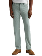 A person in AG Jeans' Everett Slim Straight Leg men's pants in light green, paired with a white shirt and brown suede shoes, stands against a plain background.