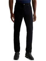 A person wearing AG Jeans Everett Sueded Slim Straight jeans with a dark shirt and gray sneakers stands facing forward against a white background.