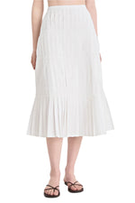 A person wearing the Litus Skirt by Merlette New York, a white pleated midi skirt, paired with black strappy flat sandals.