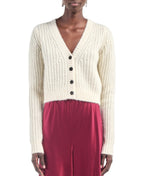 A person wearing the Maria McManus Shrunken V Cardigan, a cream-colored, cropped V-neck sweater, layered over a red satin dress and standing against a white background.