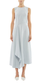 A woman wears the Adam Lippes Lynn Silk Crepe Dress, a sleeveless light blue midi design with an asymmetric draped panel, paired with white pointed-toe shoes and standing against a plain background.