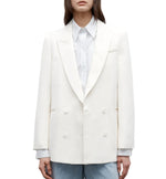 A woman wears the TWP Tabitha Blazer—a white cotton-linen, double-breasted jacket—over a light collared shirt and blue jeans, standing against a white background.