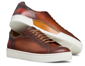 The Amadeo Sneaker by Magnanni features brown leather and white rubber soles; one stands upright while the other lies on its side, highlighting the signature Artesano sole and unique tread pattern.