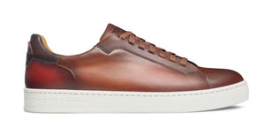The Magnanni Amadeo Sneaker is a brown leather low-top with matching laces and a white Artesano sole, displayed in profile against a white background.