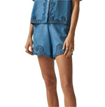 A person poses against a plain background wearing Carolina K's Roma Shorts—high-waisted with dark blue wave-like embroidery and a stylish front pleat—paired with a blue short-sleeve shirt.