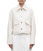 A person stands against a plain background wearing the Cropped Swing Jacket by Maria McManus, featuring an oversized collar and large pockets, paired with white trousers.