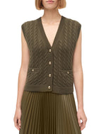 A woman wears the Simkhai Perrie Vest—an olive green, sleeveless, cable-knit cashmere vest with gold buttons—paired with a pleated skirt.