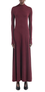 A person wearing the Maria McManus Rib Godet Dress—long-sleeved, ankle-length merlot with a fitted bodice and high neckline—and black shoes, stands against a white background.