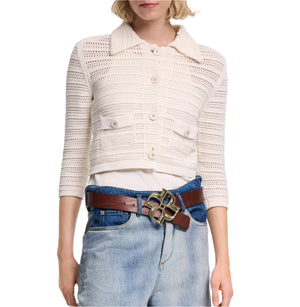 A woman wears the Dorothee Schumacher Precise Pointelle Jacket—a white crocheted button-up cardigan with graphic pointelle details—paired with a white top, blue jeans, and a brown belt featuring a large decorative buckle.