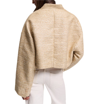 A person wears Dorothee Schumacher’s Shiny Coolness Jacket—a textured, light gold Lurex-tweed piece with a boxy silhouette and long sleeves—paired with off-white pants, shown from the back against a white background.