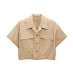 The Poplin Power Cropped Blouse by Dorothee Schumacher is a beige cotton poplin top featuring short sleeves, an open collar, two large chest pockets, and an elastic hem for a cropped fit.