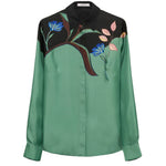 The Seductive Bloom Blouse by Dorothee Schumacher is a silk twill, long-sleeve, button-up top featuring a green body, black shoulders, and a floral print with blue flowers and pink leaves on the upper front.