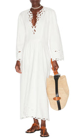 Someone is wearing the Tianny Dress by Isabel Marant, a long white cotton garment with openwork embroidery and cut-out patterns, paired with a straw tote that has leather straps and brown sandals.