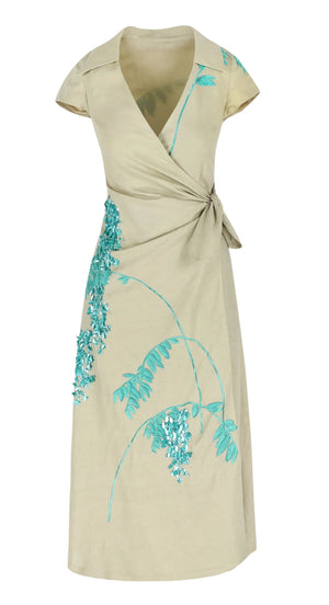 The Mara Embroidered Dress by Bernadette is a beige wrap dress with short sleeves, a side knot, and blue floral embroidery on the shoulder and skirt, shown against a plain white background.