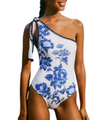 A woman models the Blue Floral Cascade One Piece by Farm Rio, featuring a white one-shoulder design with blue floral patterns and a bow detail on the left strap, set against a white background.
