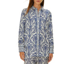 A woman wears the Farm Rio Blue Tropical Cameo Off-White Pajama Shirt, a long-sleeve, button-up top with a blue and white floral print made from LENZING™ ECOVERO™, standing against a plain white background.
