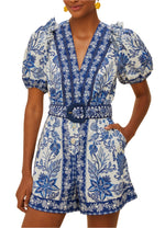 A person wears the Shell Garden Off White Romper by Farm Rio, featuring tropical florals, puffed short sleeves, a V-neckline, and a matching belted waist.