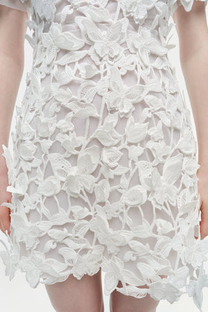 A person wearing the Simkhai Darla Mini Dress, featuring a sheer mesh base and intricate 3D floral guipure embroidery, viewed from the front.