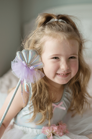 A young girl beams, holding a Great Pretenders Mermaid Wand with a seashell tip and purple tulle, embodying the under-the-sea theme. Her light-colored dress, accented with a cute pink bow, makes her ready for any enchanting kids' adventure.