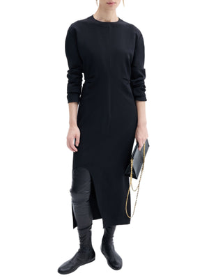 A person wearing CO's minimalist black Keyhole Dress with long sleeves, paired with black boots and holding a black clutch with a gold chain, stands against a plain white background.