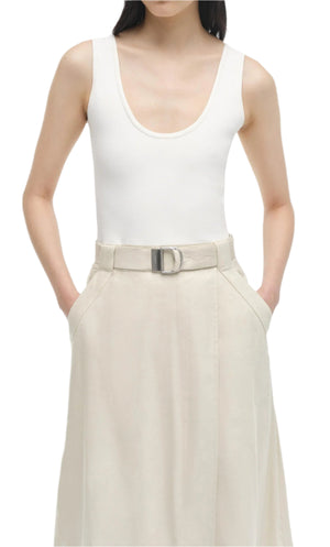 A woman wearing the Simkhai Camden Tank Midi Dress in sleeveless white linen knit, featuring a scoop neck and high-waisted beige belt detail, stands with her hands in the dress pockets.