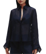 A woman wears the TWP Boyfriend Silk Organza Shirt, featuring sheer striped fabric, long sleeves with wide cuffs, and a relaxed fit, paired with dark pants.