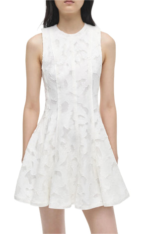 Wearing Simkhai's Sylvia Mini Dress, a sleeveless mini featuring a textured floral pattern like Summer Jacquard, the person stands with hands in pockets against a plain backdrop.